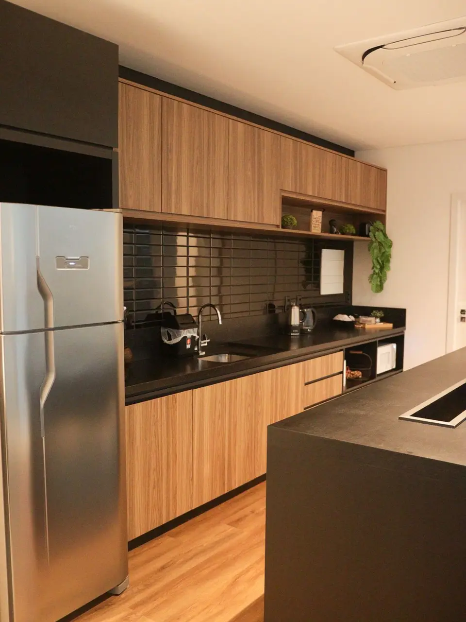 Cozinha do escritório com armários de madeira, bancada preta e geladeira inox.