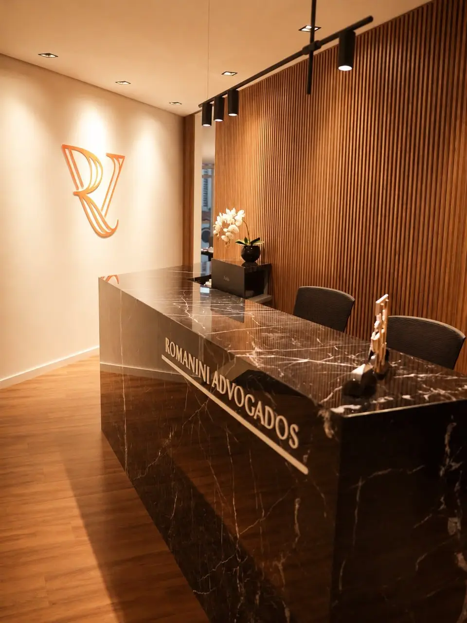 Recepção com o monograma RV laranja na parede e o letreiro Romanini Advogados no balcão de mármore preto.